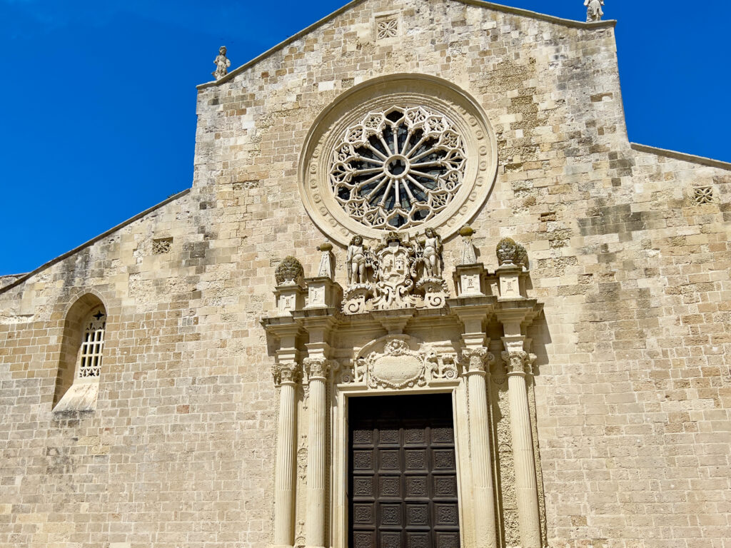 Otranto Cathedral (Basilica di Santa Maria Annunziata)

The cathedral is built on the site of earlier Messapian, Roman, and early Christian structures. Its façade, dating back to 1088, rises prominently on Piazza Basilica and stands as one of the most significant examples of Apulian Romanesque architecture. A 15th-century rose window, displaying Gothic-Arabic design, adorns the center of the façade, overlooking a 17th-century portal. The bell tower, coeval with the Cathedral, stands in the northeastern corner of Piazza Basilica and takes on a square plan spanning two stories.

Photo by the Puglia Guys, Puglia ambassadors. PugliaGuys.com